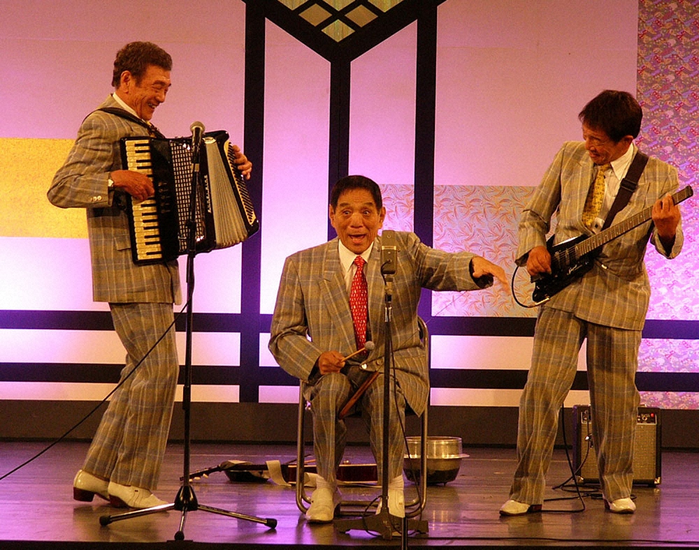 2012年6月、手術からの復帰舞台でおなじみのノコギリ芸を披露した横山ホットブラザーズの長男・アキラさん（中）に、次男・マコト（左）三男・セツオも笑顔を見せる