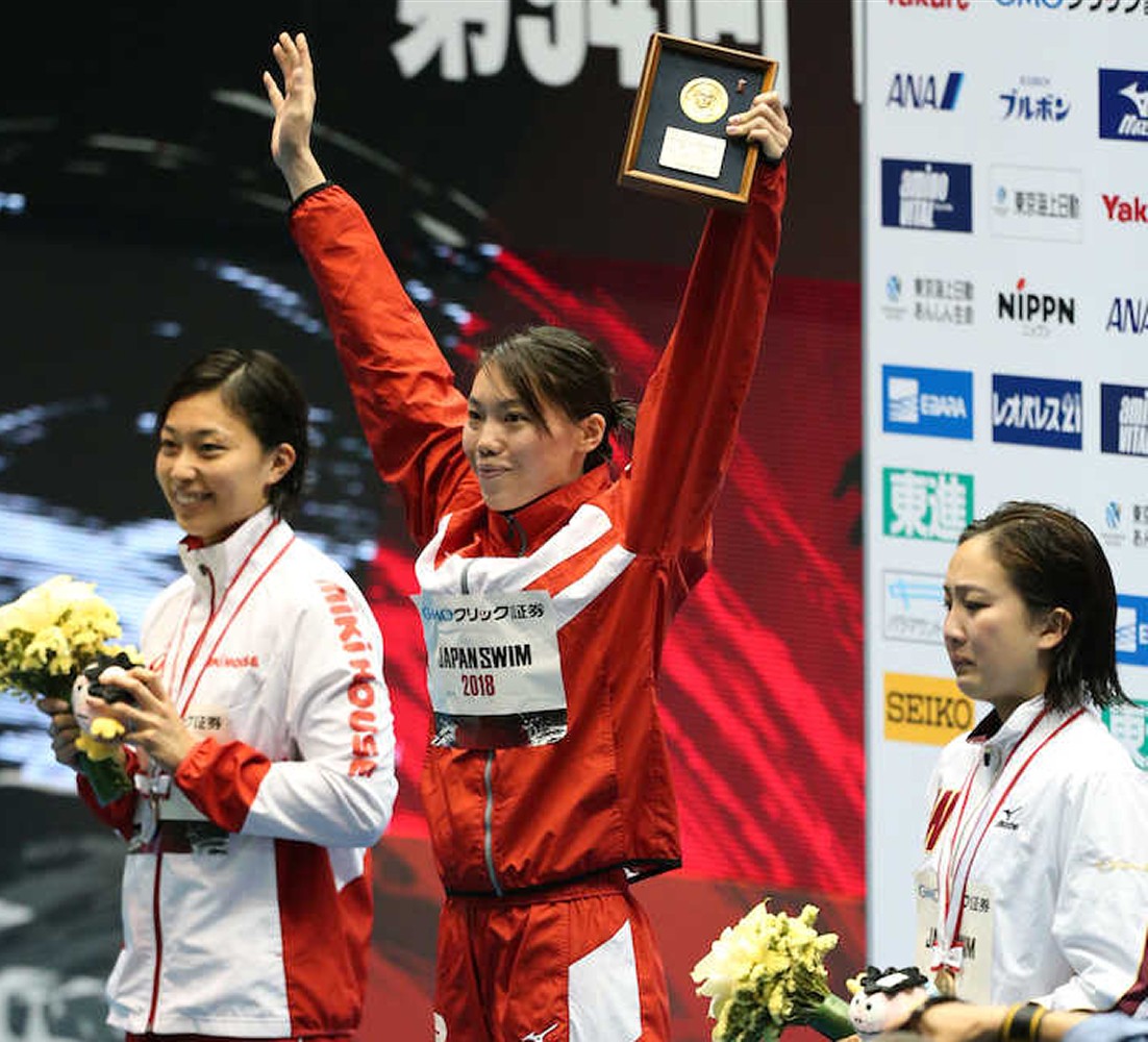 女子１００メートル平泳ぎで優勝し、表彰台で声援に応える青木（中央、左は２位の鈴木、右は３位の渡部）